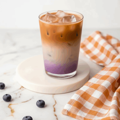 Creamy blueberry latte topped with foam and fresh blueberries in a glass mug on a wooden table